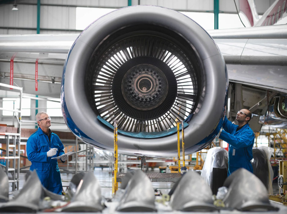 Two aviation technicians in blue coveralls inspect a jet engine inside a hangar, front view of the turbine impeller visible.