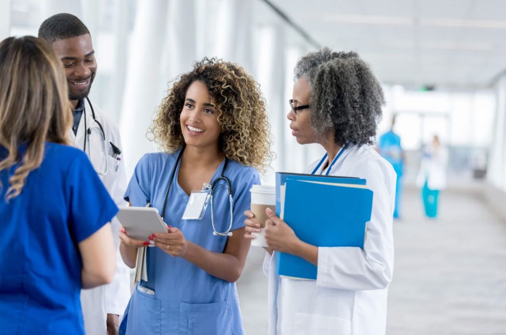 A diverse team of doctors and nurses in a hospital corridor discuss patient care, with a nurse showing a tablet to colleagues.