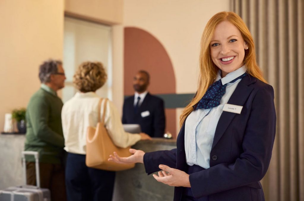 Smiling hotel front desk attendant in a navy blazer greets guests at a reception desk in a lobby area.