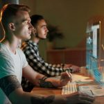 Two young men at a desk using a computer with holographic data overlays in teal and blue, showing charts and graphs in front of them.