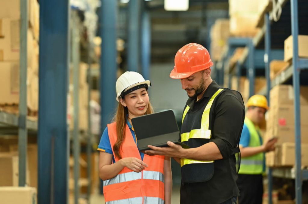 Warehouse workers review inventory on a tablet together in a storage rack area, wearing safety vests and hard hats.