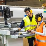 Two factory workers in high‑visibility vests and safety gear review documents at an industrial conveyor in a manufacturing facility.