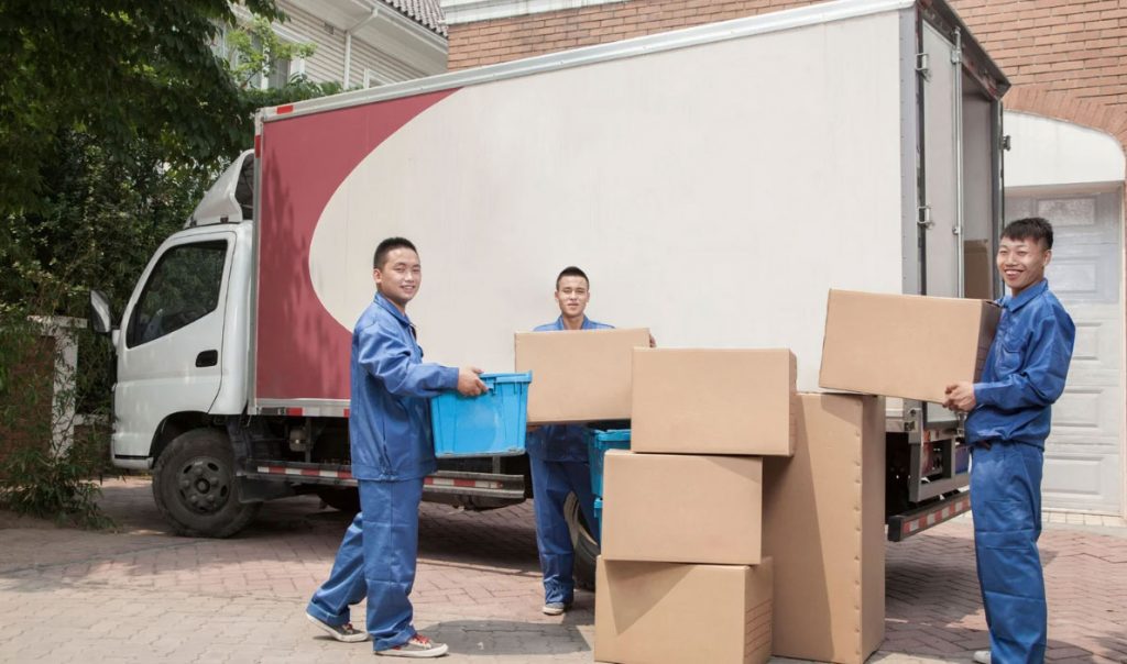 Delivery crew in blue uniforms unload cardboard boxes from a white delivery truck parked in a driveway.