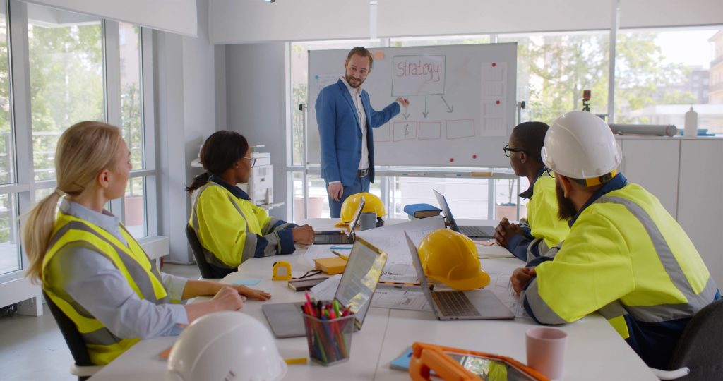 ConstructionArchitectsOnMeetingAndPresentationInBrightModernOffice - Language Training Pro Team of construction workers in high-visibility jackets listen to a presenter in a blue suit as he explains a strategy on a whiteboard in a bright meeting room.
