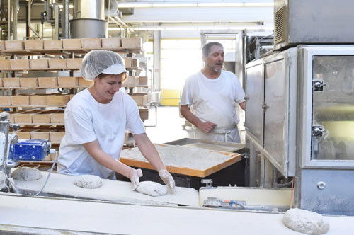 Baker in a hairnet shapes dough on a conveyor while another worker watches near an industrial oven in a bakery setting with stacked trays in the background.