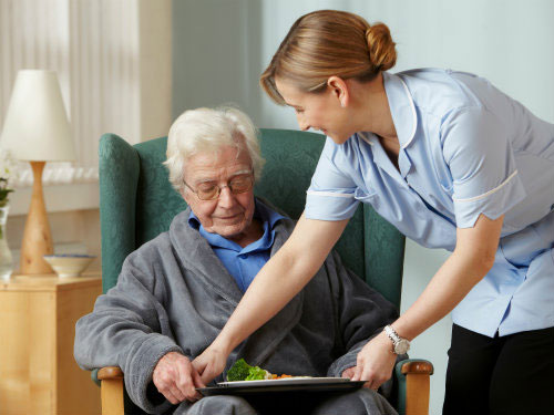 Caregiver in a blue uniform assists an elderly man in a chair, guiding him to eat his meal on a plate at home.
