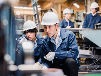 Group of factory workers in white helmets and blue coveralls inspecting machinery in a manufacturing plant.