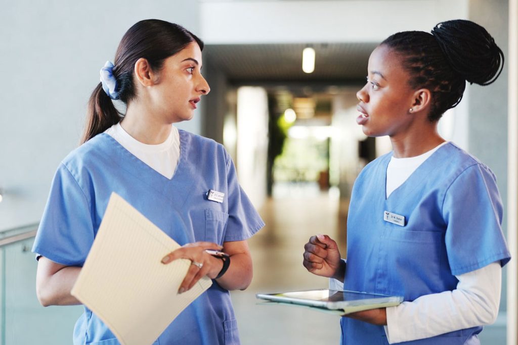 3-English-training-for-nurses-and-CNAs - Language Training Pro Two nurses in blue scrubs stand in a hallway, talking; one holds a yellow notebook while the other carries a tablet.