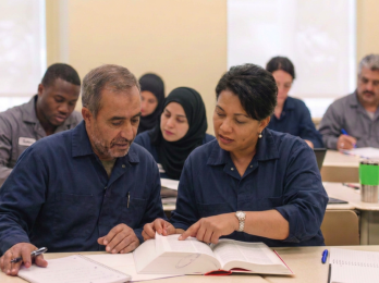 Two adults in blue uniforms read and discuss an open book at a classroom desk, with classmates in the background.