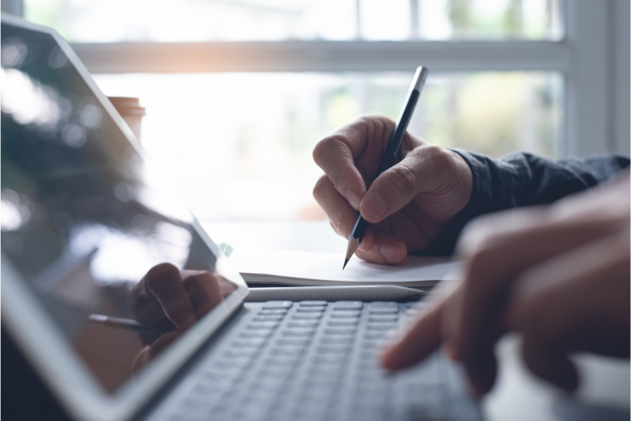 Close-up of a person writing with a pencil on a notebook, with a laptop and tablet nearby.