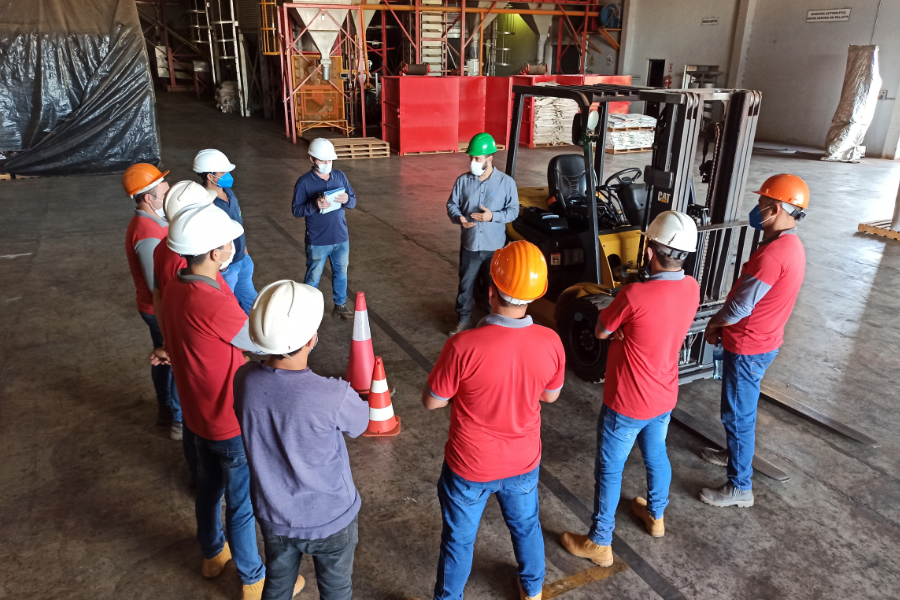 Safety training manufacturing - Language Training Pro Group of workers in a warehouse standing in a circle for a safety briefing near a forklift and traffic cones, all wearing hard hats and masks.