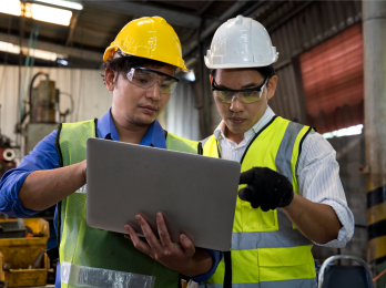 Two construction workers in hard hats and high‑visibility vests inspect data on a laptop in an industrial setting