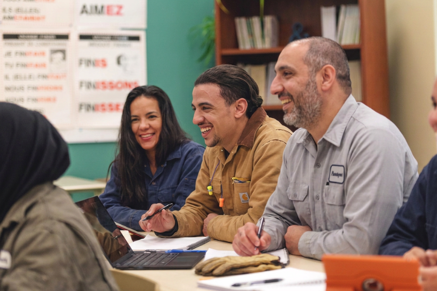 Immigrant manufacturing workers laughing together during a workplace English language class, part of an employee retention and career pathway program.