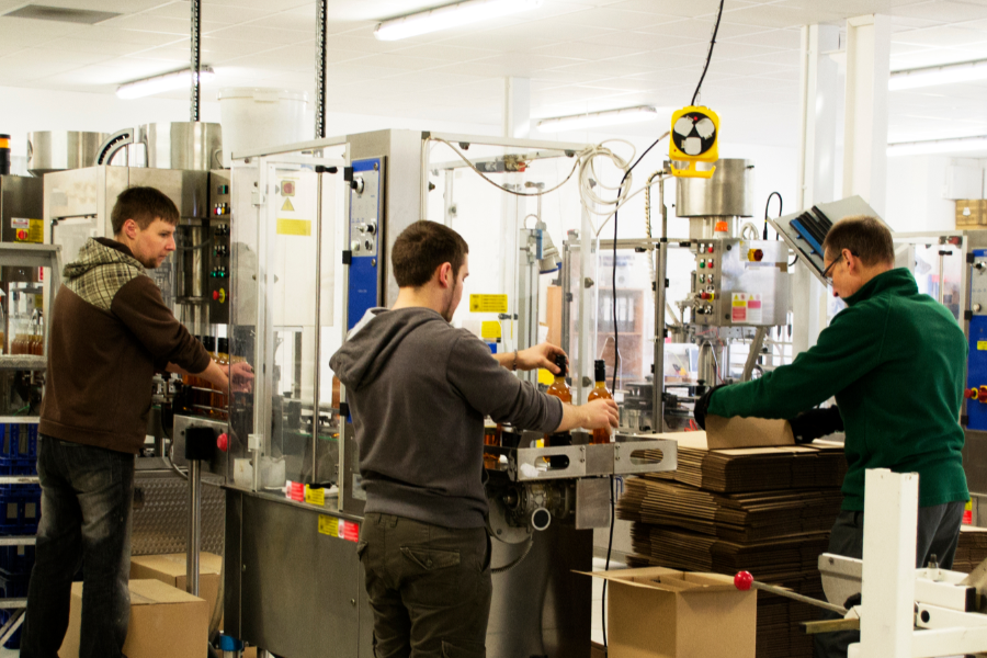 Three workers operate a bottling and packing line in a factory, filling bottles and boxing them.