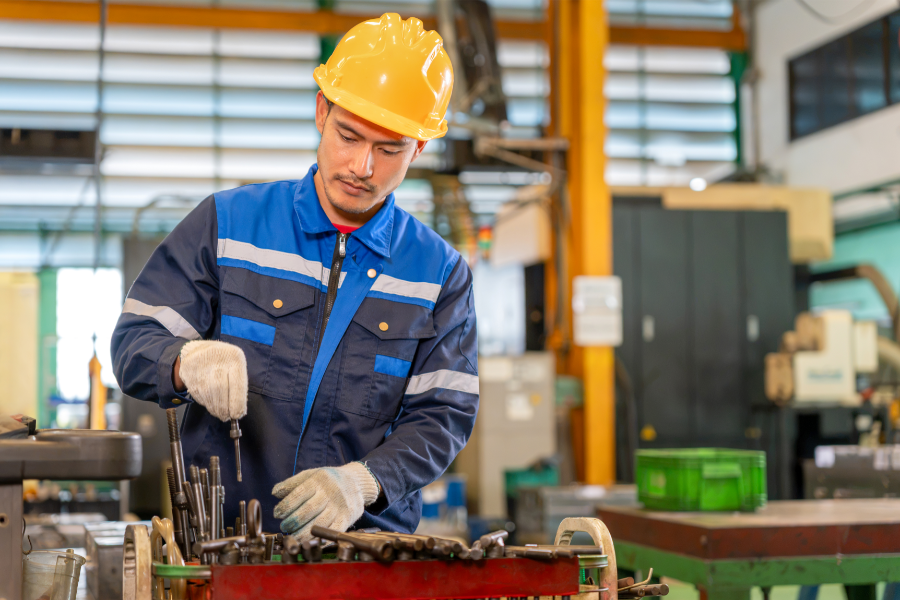 Male factory worker in a blue uniform and yellow hard hat assembling tools at a workbench, wearing gloves in an industrial setting
