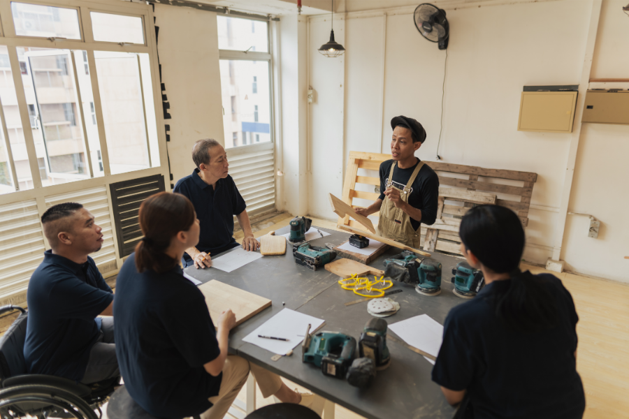 Instructor in overalls demonstrates a woodworking technique to a small group around a table with power tools in a bright workshop.