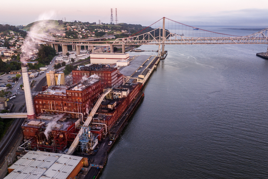Aerial view of a waterfront industrial complex with smokestacks and red brick buildings beside a river, with a large bridge in the background.