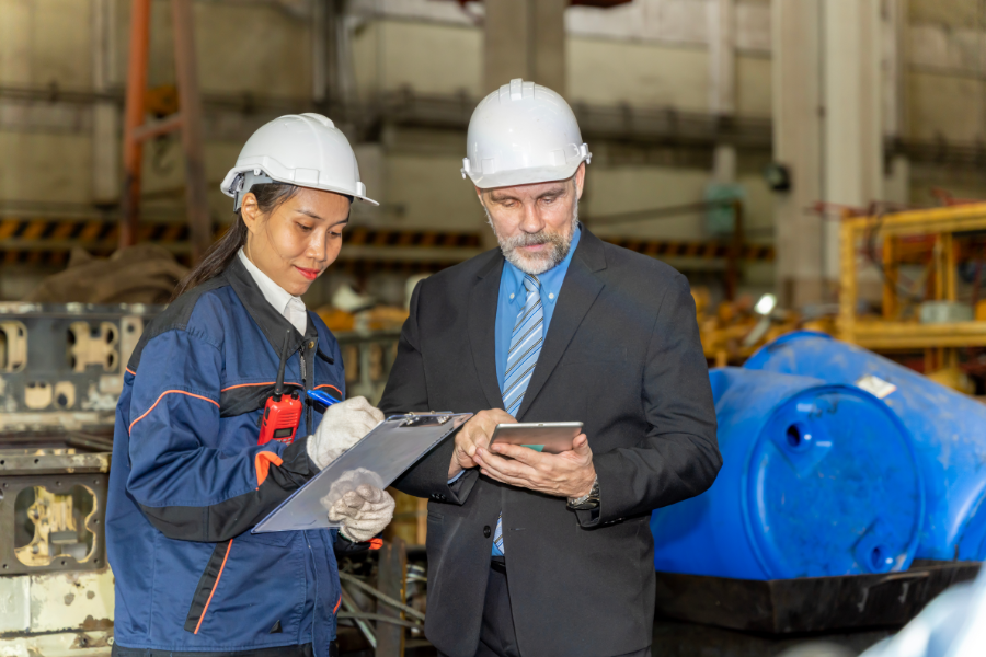 Two workers in hard hats review plans: a woman with a clipboard and a man with a tablet in a factory setting. online English training course
