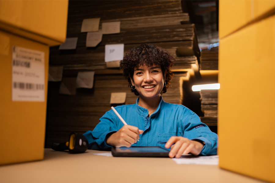Smiling woman in a blue shirt writes on a tablet at a desk surrounded by cardboard boxes in a warehouse setting.