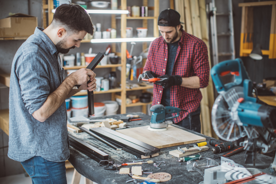 Two craftsmen in a cluttered workshop measure and shape wood at a workbench with power tools nearby